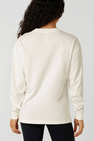 A person with long, wavy dark hair is shown from behind, wearing a plain white Lorna Jane Country Club Long Sleeve T-Shirt and black pants against a light gray background.