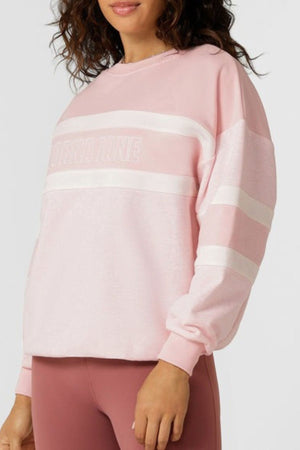 A woman wears the Lorna Jane Dreamer Oversized Sweat in light pink with white stripes and matching leggings. The cotton blend terry sweatshirt has a relaxed fit and subtle chest text, set against a plain background.