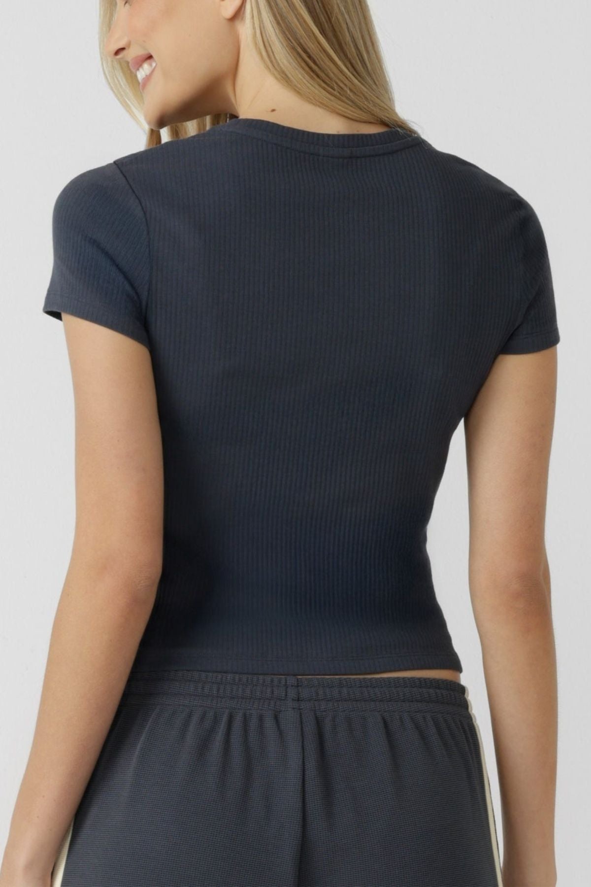 Smiling and showcasing her everyday style against a plain light background, a woman with long blonde hair wears the fitted, short-sleeve Lorna Jane Everyday Rib T-Shirt in a dark hue, paired with matching soft cotton-blend pants and shown from the back.