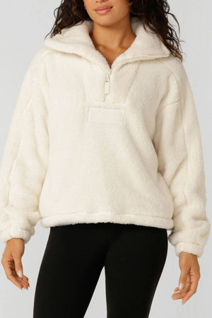 A woman wears the Lorna Jane Everyday Teddy Quarter Zip Sweat, a cozy cream sherpa fleece pullover with a high collar, long sleeves, and an oversized fit, styled with black leggings.