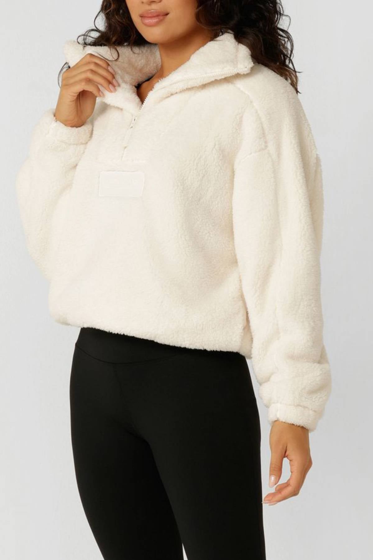 A woman wears the Lorna Jane Everyday Teddy Quarter Zip Sweat, featuring a cozy oversized fit and 100% polyester, with black leggings. She stands against a plain light background, her face partially out of frame.