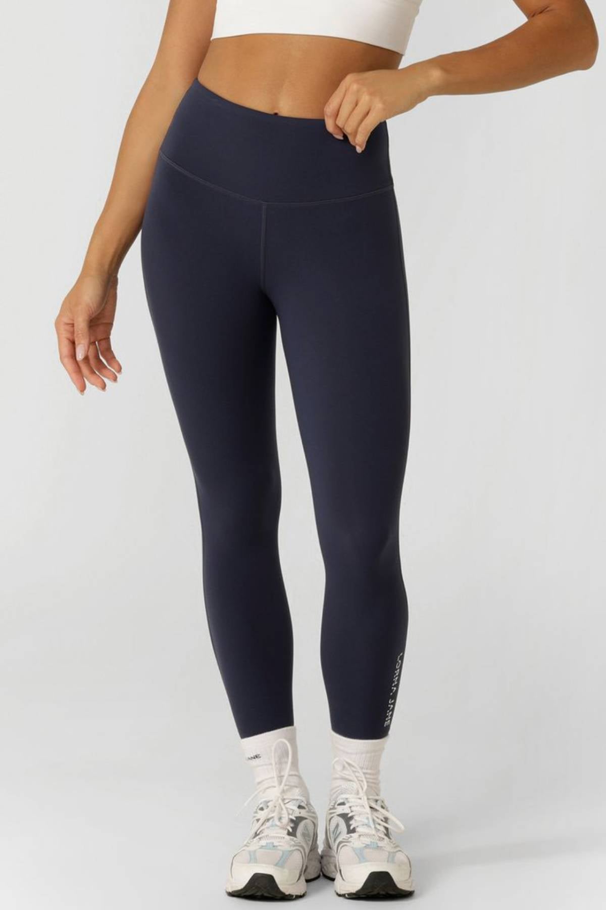 A person wearing a white crop top, navy blue Lorna Jane Lotus No Chafe Phone Pocket Ankle Biter Leggings, white socks, and white athletic shoes stands against a plain light gray background. Their face is not visible.