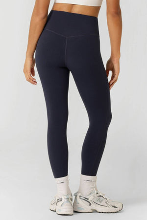 Someone stands with their back to the camera in dark blue Lorna Jane Lotus No Chafe Phone Pocket Ankle Biter Leggings, a white sports bra, white socks, and gray New Balance sneakers against a plain background, highlighting stylish activewear.