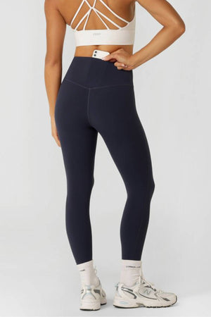 A woman faces away in a white sports bra, white socks, and sneakers, wearing Lorna Jane Lotus No Chafe Phone Pocket Ankle Biter Leggings—modern activewear with a small pocket against a plain light background.