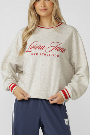 A woman wears the Lorna Jane Golden Era Sweat, an oversized light gray cotton blend terry sweatshirt with a red embroidered logo, red-striped cuffs and collar, paired with navy blue athletic pants against a plain background.