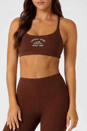 A woman wears a matching brown Lorna Jane Limited Edition Lotus Crop Sports Bra and high-waisted leggings set with a strappy back and white "Lorna Jane Luxe Athletic Since 1989" text, standing with one hand by her side.