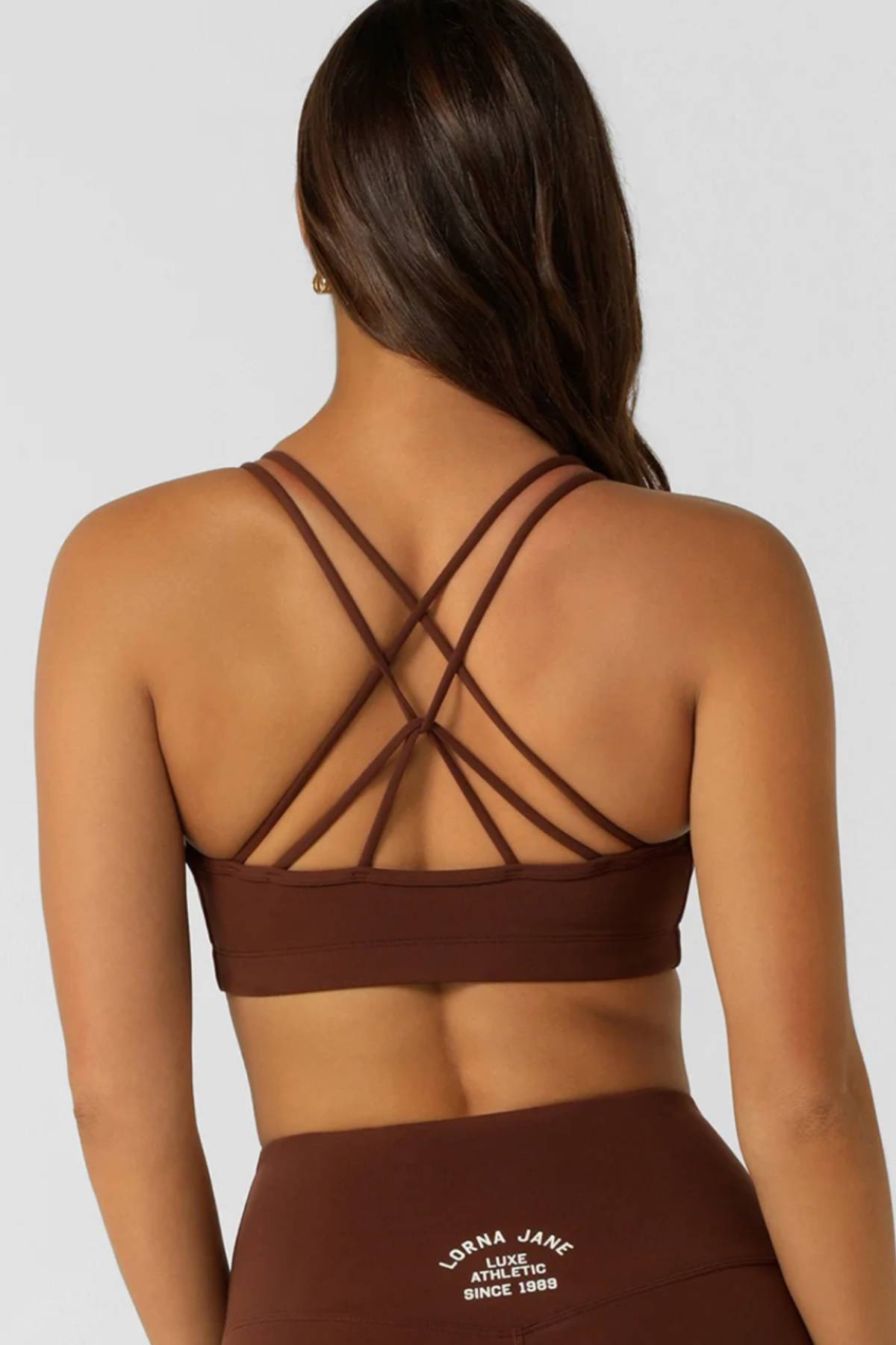 A woman with long brown hair is seen from the back, wearing the Lorna Jane Limited Edition Lotus Crop Sports Bra and matching high-waisted leggings featuring "Lorna Jane Athletic Since 1989" printed on them.
