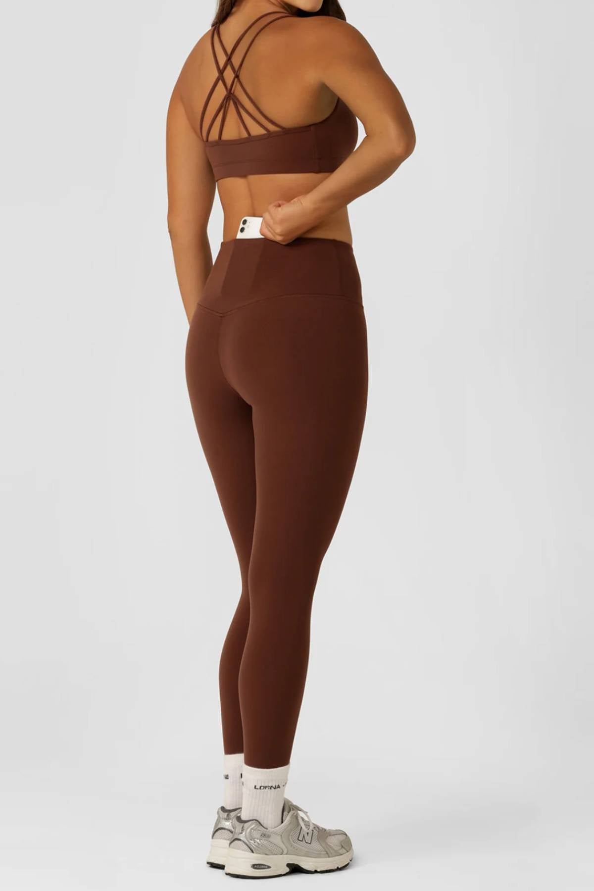 A woman stands sideways on a plain light background, turning her head. She wears a brown sports bra, white socks, gray sneakers, and holds the high-rise waistband of her Lorna Jane Limited Edition Lotus Full Length Leggings.