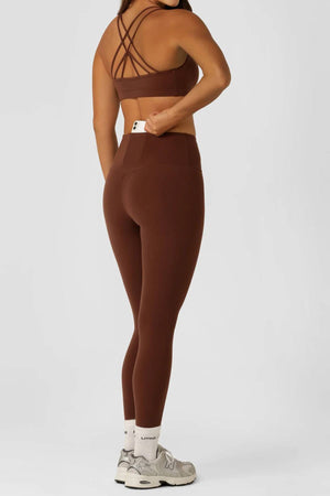 A woman stands sideways on a plain light background, turning her head. She wears a brown sports bra, white socks, gray sneakers, and holds the high-rise waistband of her Lorna Jane Limited Edition Lotus Full Length Leggings.