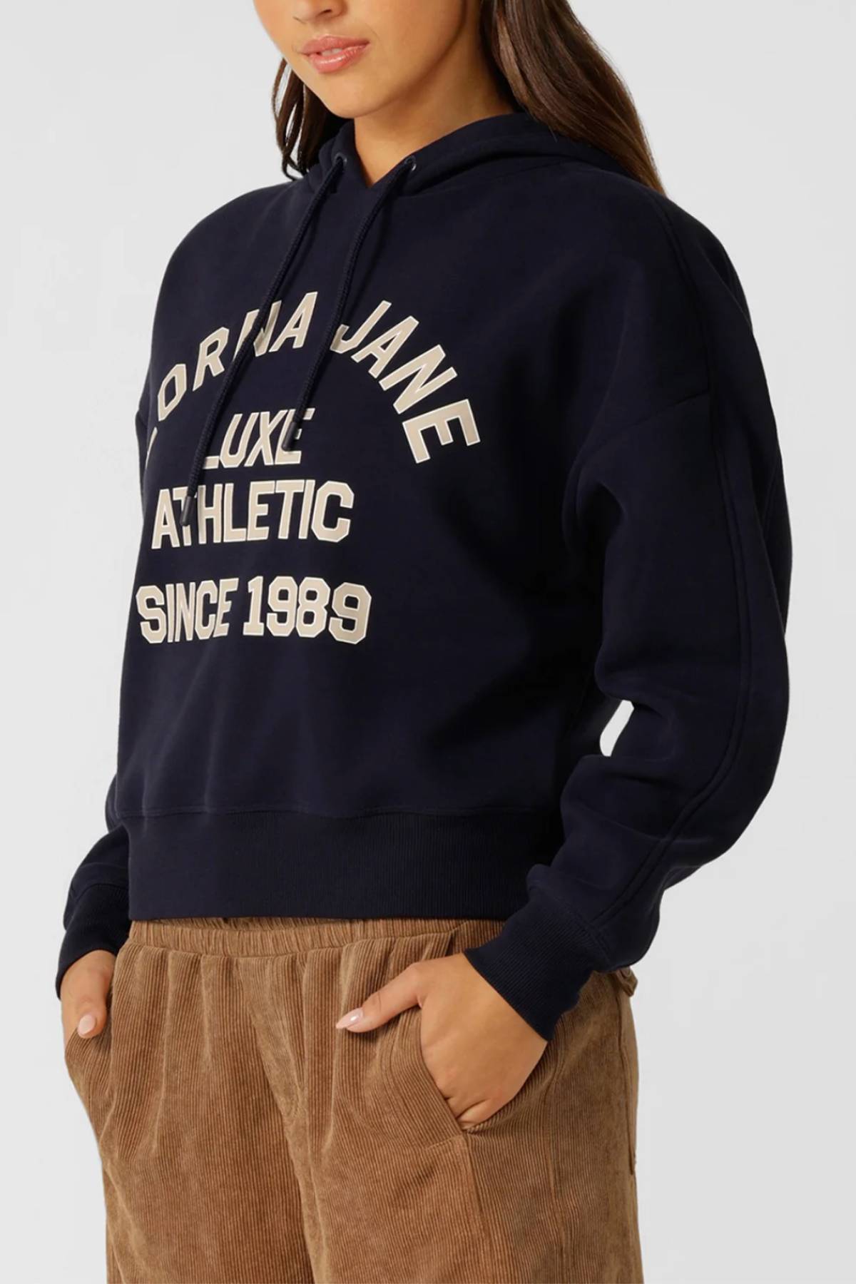 Woman wears the Lorna Jane Limited Edition Lotus Hoodie in brushed fleece, with "Lorna Jane Luxe Athletic Since 1989" on the front, paired with brown corduroy pants and hands in pockets against a plain background.