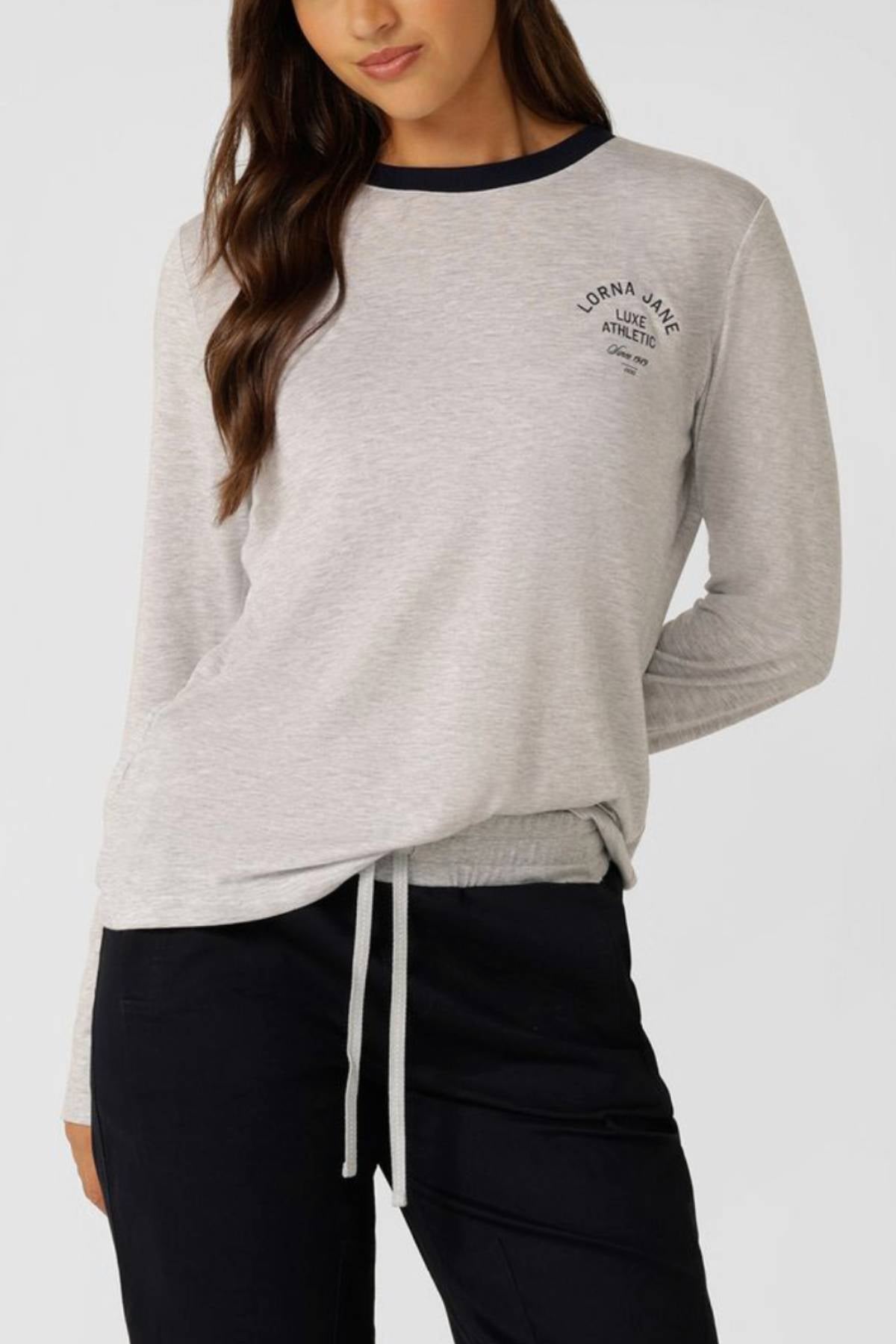 A woman wears the Lorna Jane Limited Edition Lotus Long Sleeve Top in light grey with a dark collar and chest text, paired with dark pants. She stands against a plain background with one hand in her pocket.