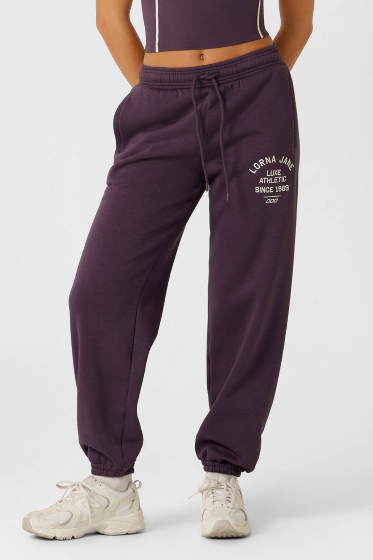 A person in Lorna Jane Limited Edition Lotus Sweatpants with "Athletic Since 1989" on the left thigh, a matching crop top, and white sneakers stands against a plain light backdrop.