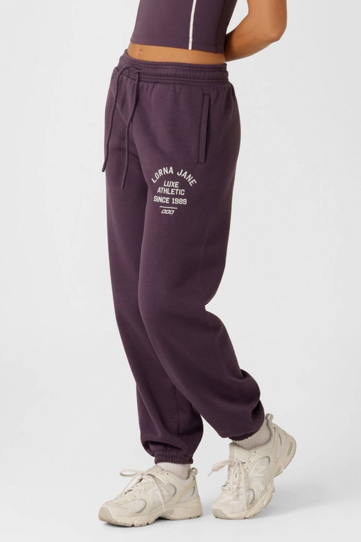 A person wearing a sleeveless fitted top and dark purple Lorna Jane Limited Edition Lotus Sweatpants with "Lorna Jane Luxe Athletic Since 1998" on the left thigh, paired with white sneakers, stands against a plain background.