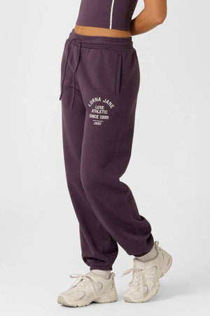 A person wearing a sleeveless fitted top and dark purple Lorna Jane Limited Edition Lotus Sweatpants with "Lorna Jane Luxe Athletic Since 1998" on the left thigh, paired with white sneakers, stands against a plain background.