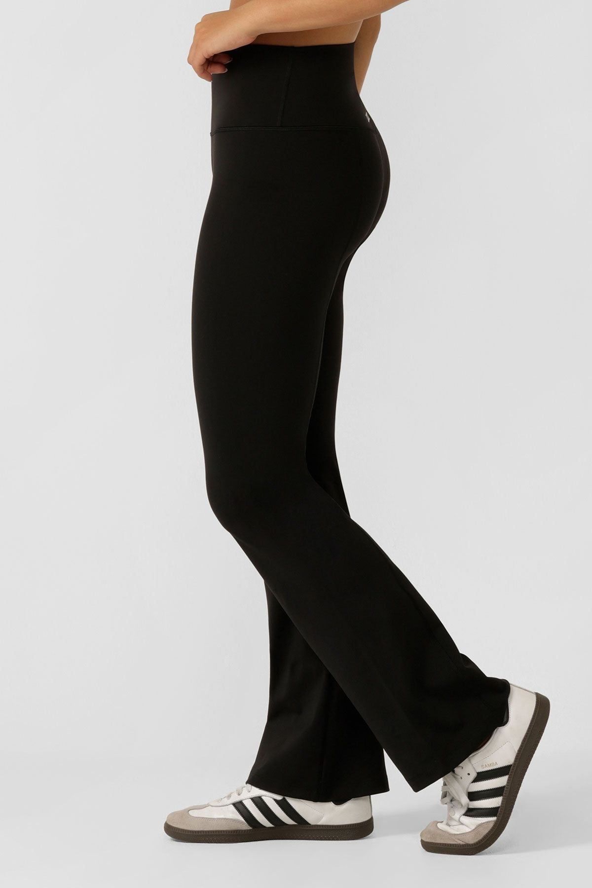 A person wearing Lorna Jane Lotus Flared Legging in black and white sneakers with black stripes stands sideways against a plain light background, showing only their lower body.