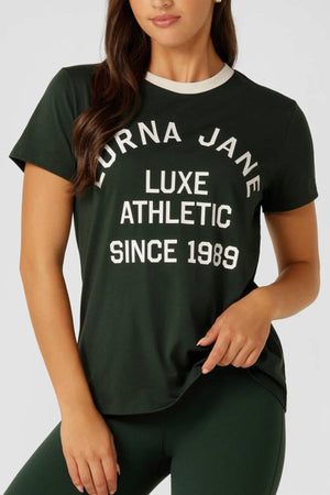 A woman wears the Lorna Jane Lotus Heritage Ringer T-Shirt in dark green with white trim and a limited edition "LORNA JANE LUXE ATHLETIC SINCE 1989" logo, paired with matching green leggings.