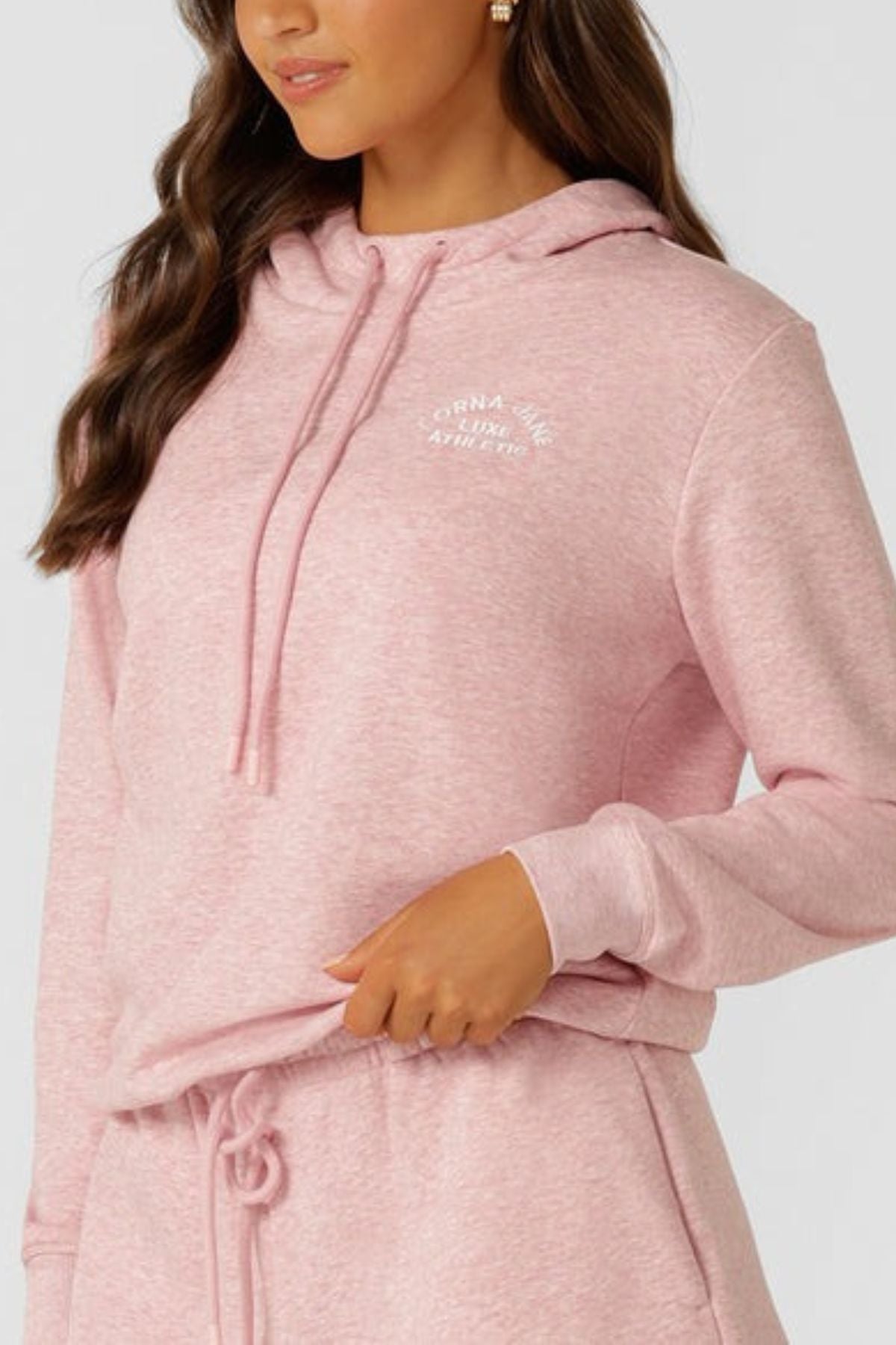 A woman in a relaxed fit, light pink Lorna Jane Lotus Hoodie made from cotton blend terry touches its waistband. The hoodie features subtle white chest text, and her long, wavy hair is down. Matching sweatpants complete the look.