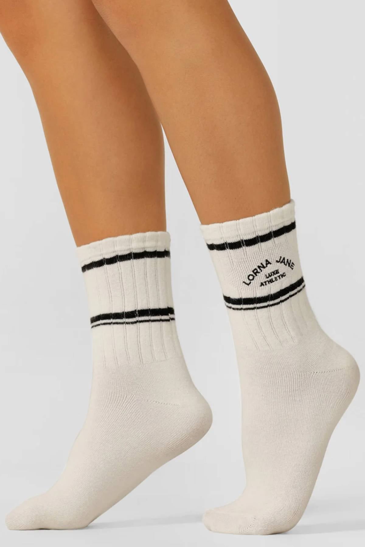 A pair of feet wearing Lorna Jane Lotus Limited Edition Quarter Crew Socksβwhite ribbed, quarter crew style with two black stripes near the top and "Lorna Jane Athletic" on the cuff. The background is plain light gray.