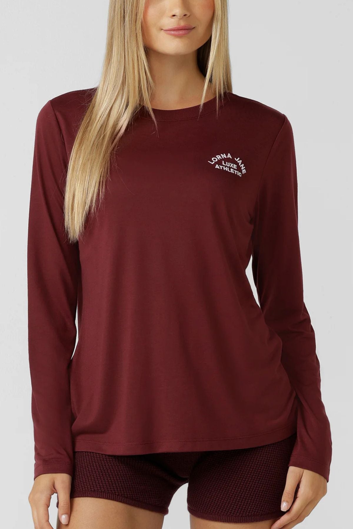 A woman in a Lorna Jane Lotus Long Sleeve T-Shirt and matching maroon shorts stands against a plain gray background, featuring a small white logo on the left chest. Her straight blonde hair complements the activewear look.