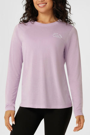 A woman wears the Lorna Jane Lotus Long Sleeve T-Shirt in light purple with a subtle white logo, styled with black leggings. She stands and smiles against a plain light background. This lightweight, breathable T-shirt is ideal for any activity.