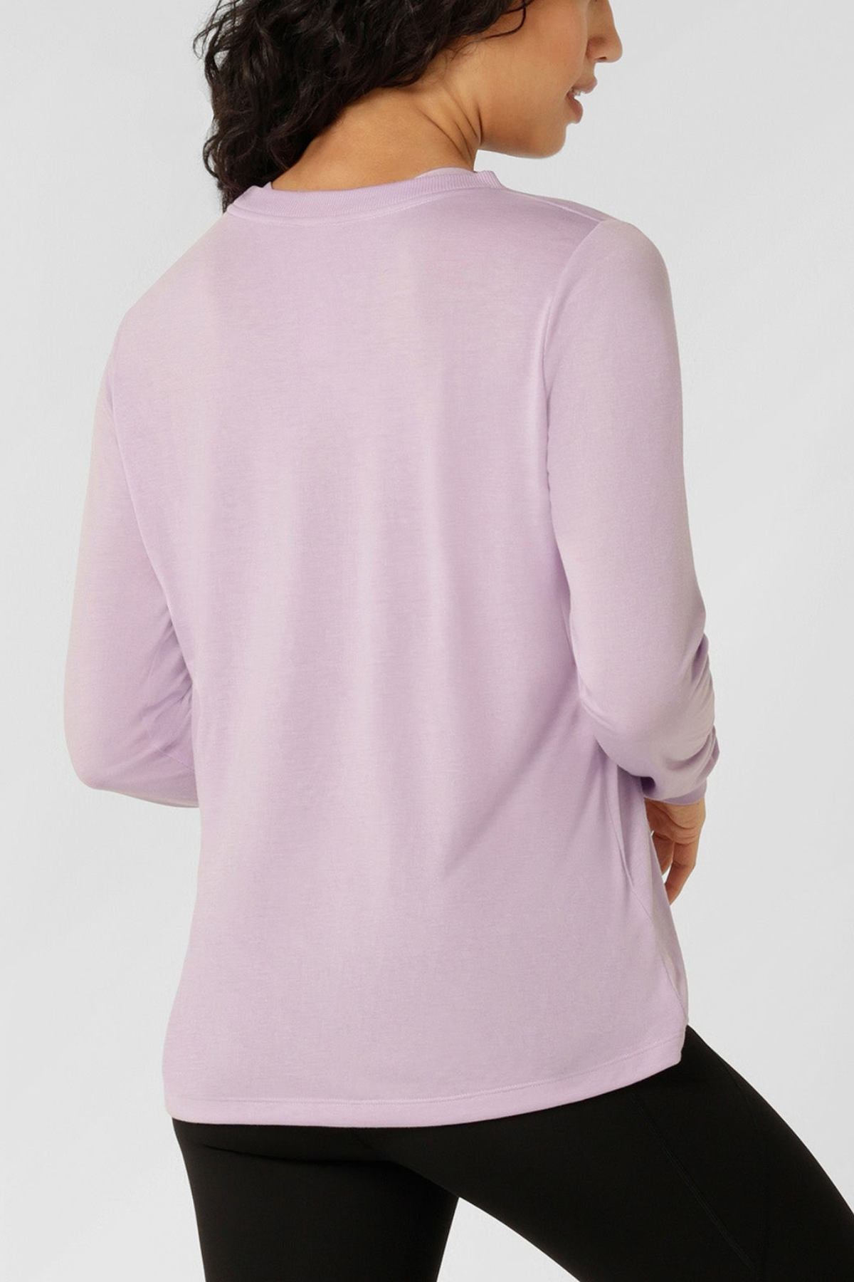 A woman with dark hair is seen from behind wearing a lightweight, breathable Lorna Jane Lotus Long Sleeve T-Shirt in light purple with black pants, standing against a plain light background.