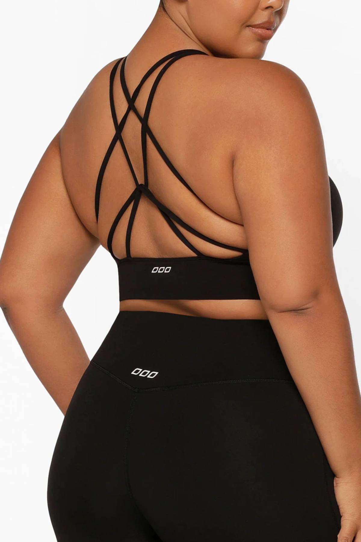 A woman is shown from behind on a white background, wearing the Lorna Jane Lotus Longline Sports Bra in black with multiple crisscross back straps and matching black leggings.