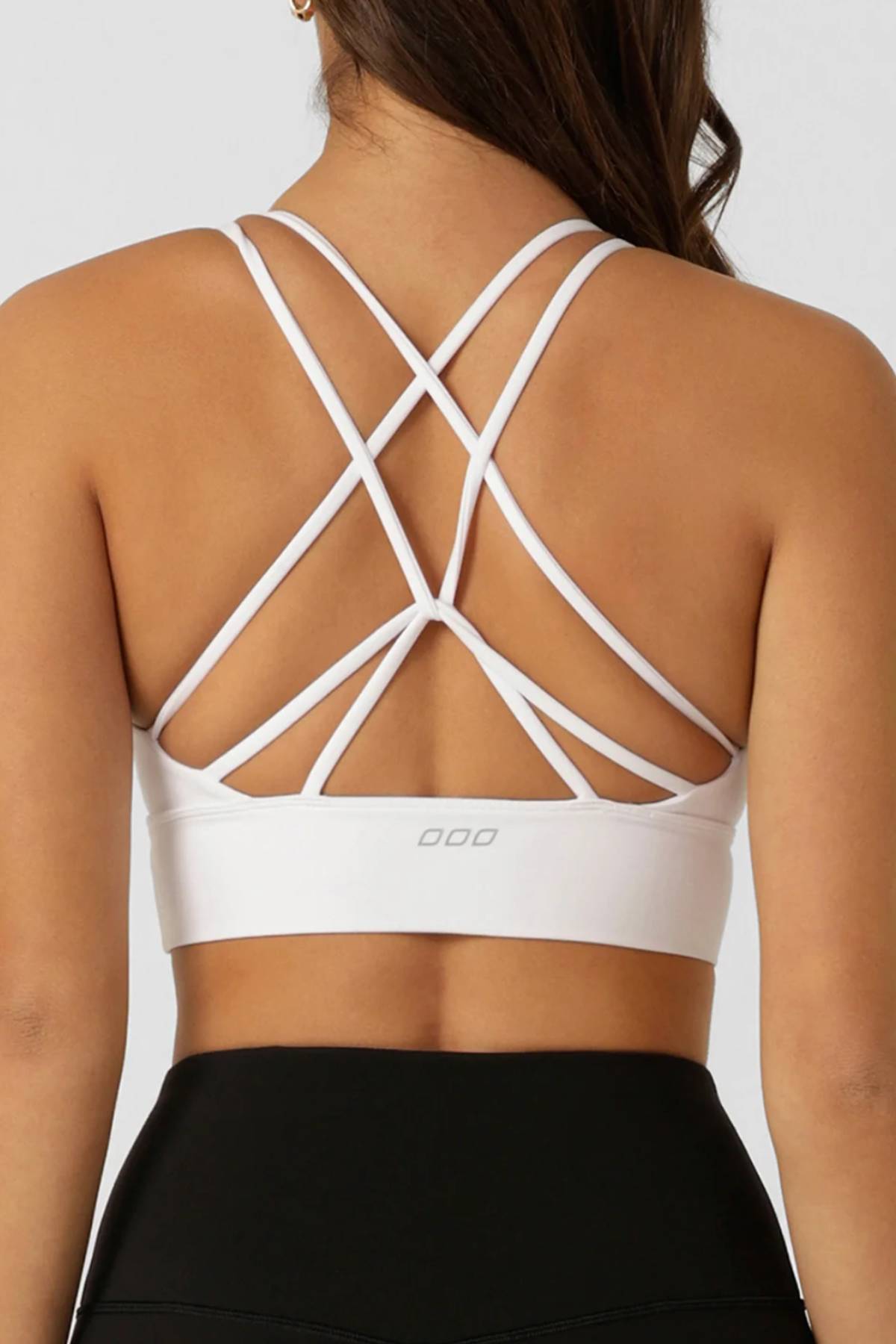 A woman wears the Lorna Jane Lotus Longline Sports Bra—white, strappy, moisture-wicking, with a crisscross back design—paired with black leggings. The image focuses on her back, showcasing the bra’s intricate straps and supportive fit.