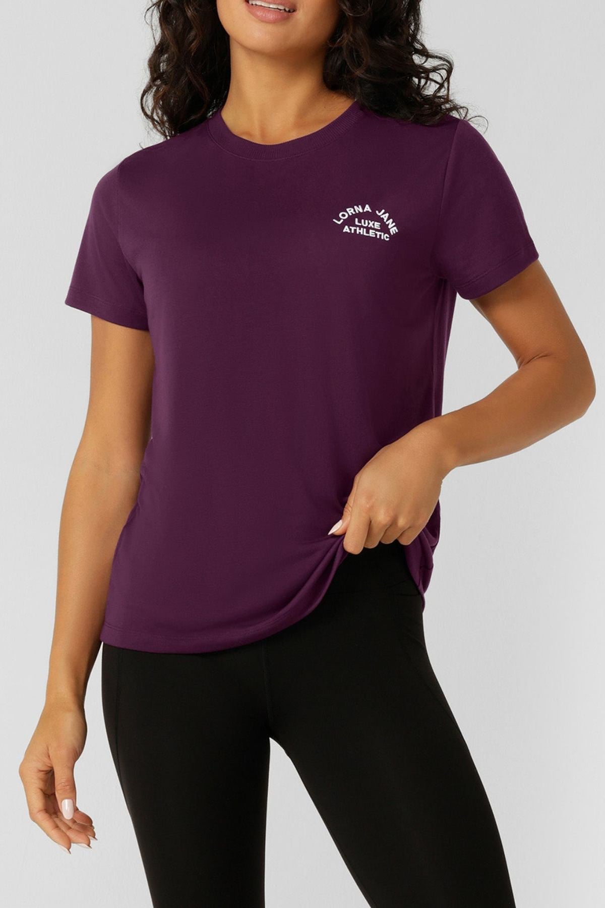 A woman wears a purple Lorna Jane Lotus T-Shirt with "Lorna Jane Athletic" in white on the chest, paired with black leggings, standing against a plain light background.