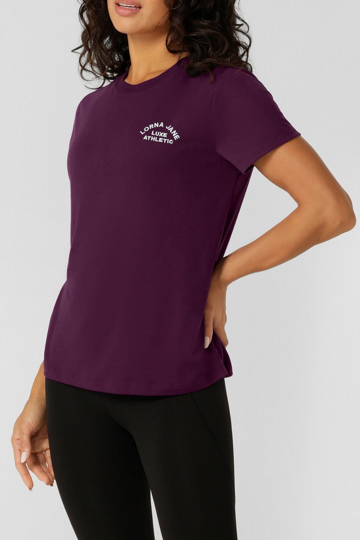 A woman models the Lorna Jane Lotus T-Shirt in dark purple, featuring "Lorna Jane Athletic" in white on the chest, paired with black leggings. She stands confidently against a light background with one hand on her hip.