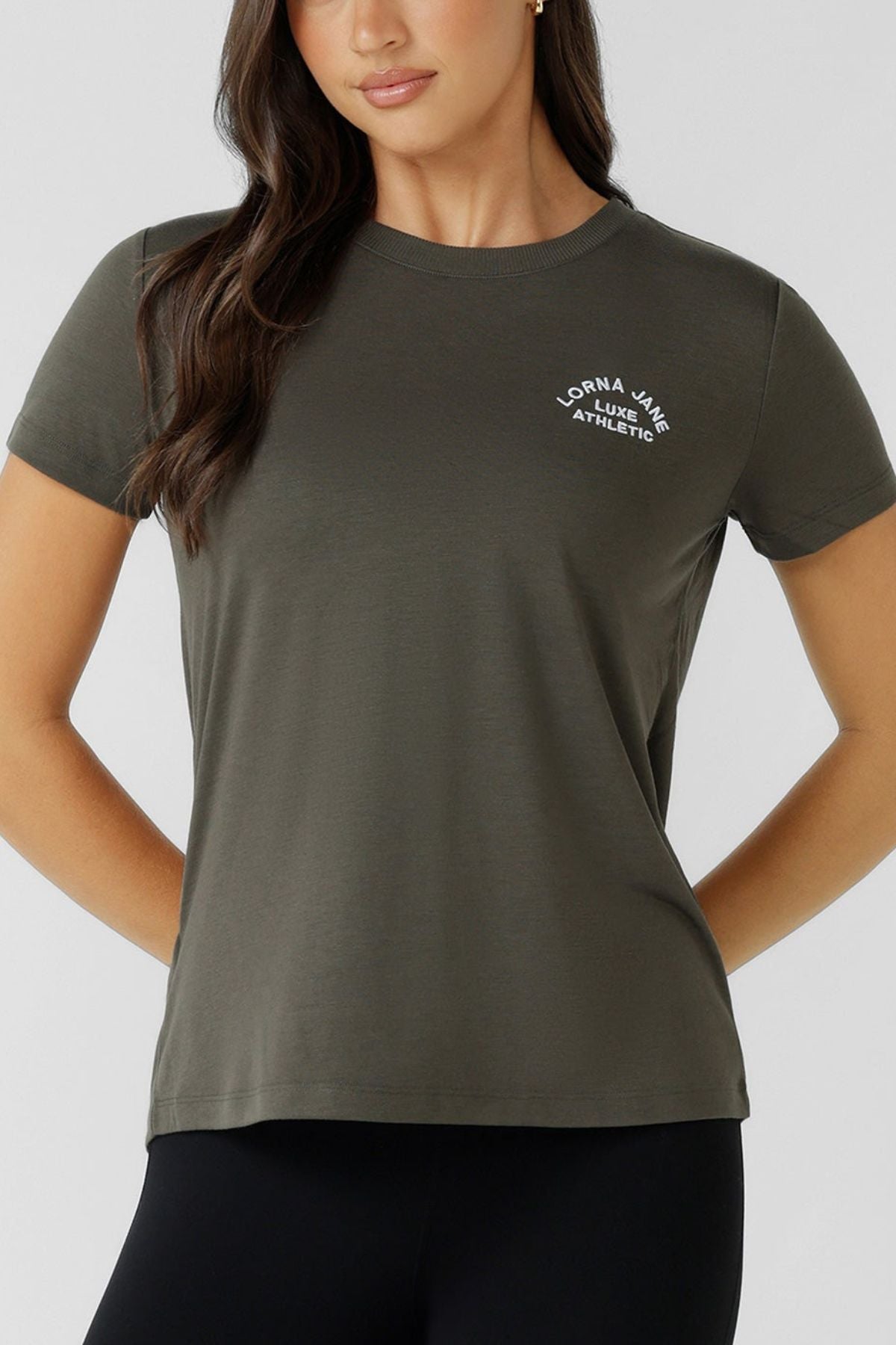 A woman confidently stands with one hand on her hip, wearing a Lorna Jane Lotus T-Shirt—an olive green women’s crew neck with “Lorna Jane Athletic” in white on the left chest—paired with black leggings.