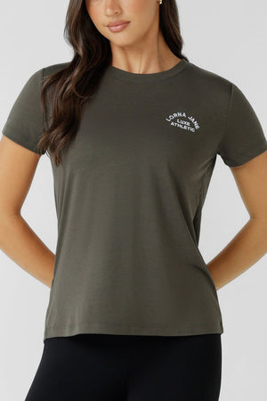A woman confidently stands with one hand on her hip, wearing a Lorna Jane Lotus T-Shirt—an olive green women’s crew neck with “Lorna Jane Athletic” in white on the left chest—paired with black leggings.