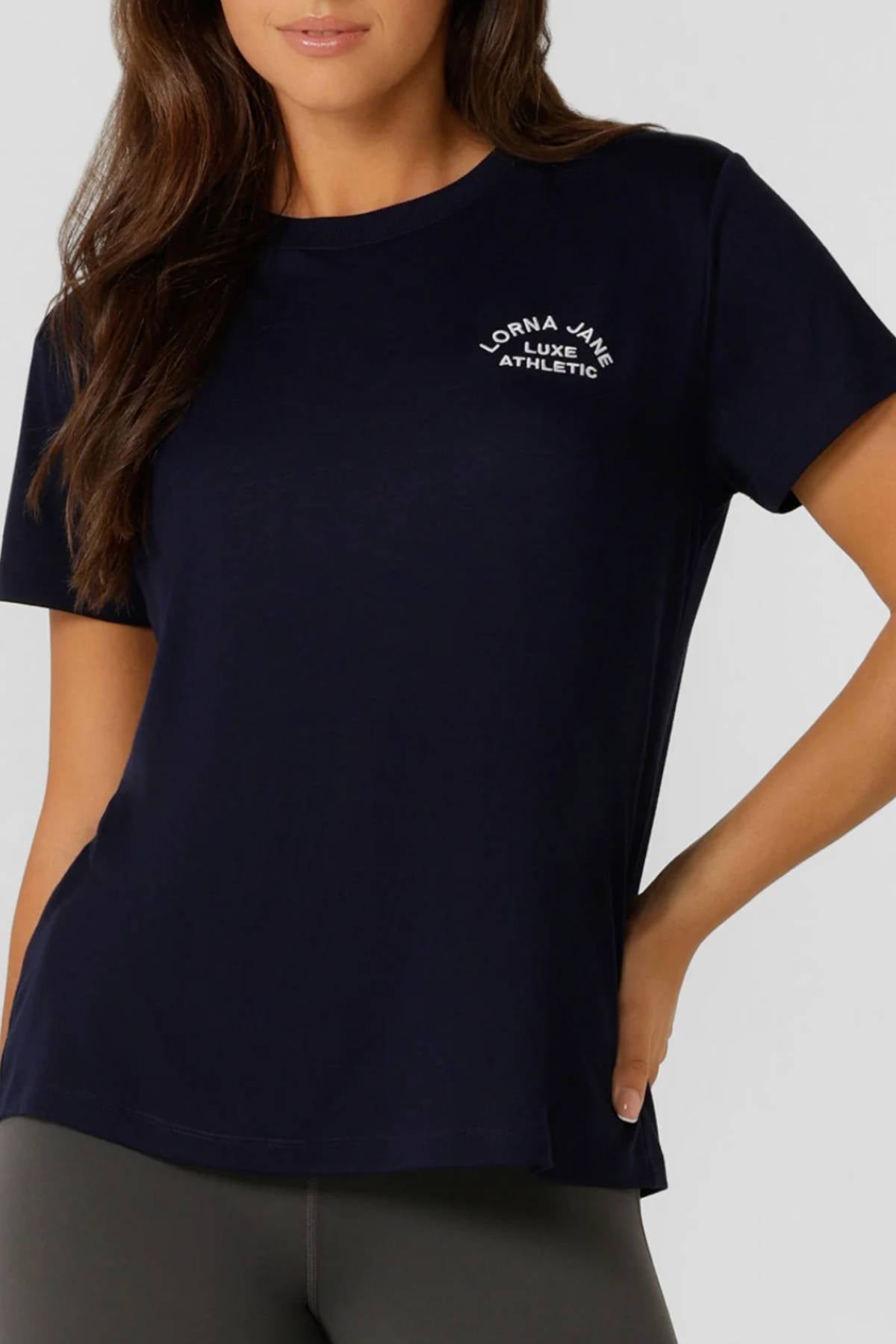 A woman models the Lorna Jane Lotus T-Shirt in navy blue with "Lorna Jane Luxe Athletic" on the chest, paired with dark leggings—ideal activewear for women who value both style and performance.