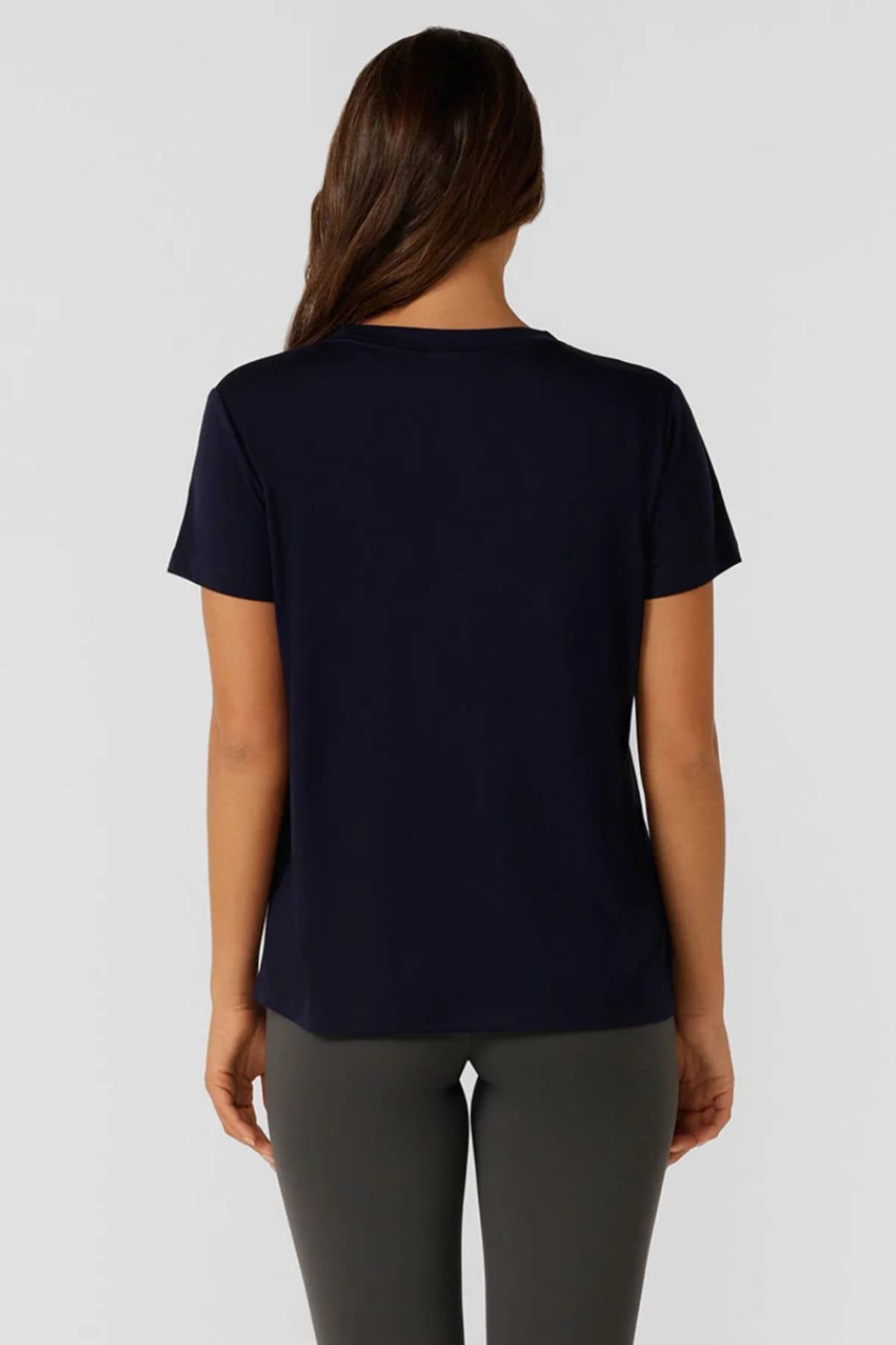 A woman with long brown hair, facing away, wears a short-sleeve dark navy Lorna Jane Lotus T-Shirt and gray pants—ideal women's activewear for an active lifestyle—set against a plain light background.