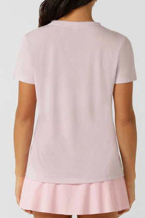 A woman is seen from the back in a light pink Lorna Jane Lotus T-Shirt with an embroidered logo, paired with a pastel pink pleated skirt against a plain, neutral background.
