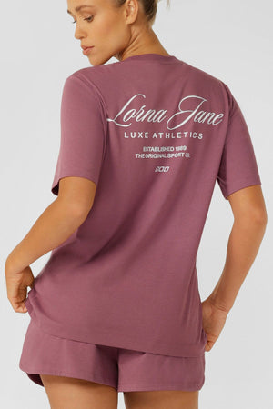 A woman faces away, wearing a mauve Lorna Jane Luxe Athletic Relaxed T-Shirt and matching shorts. The back of her shirt displays "Lorna Jane Luxe Athletics Established 1989 The Original Sport Co." in white text.