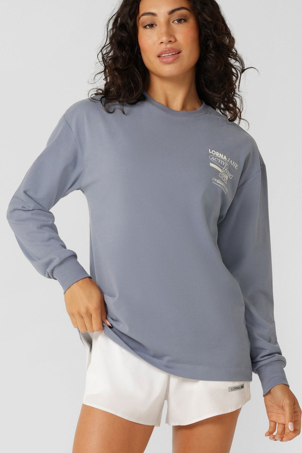 A woman with wavy dark hair wears a loose gray cotton Lorna Jane Momentum Plush Long Sleeve T-Shirt with white chest text and white athletic shorts, standing against a plain light background.