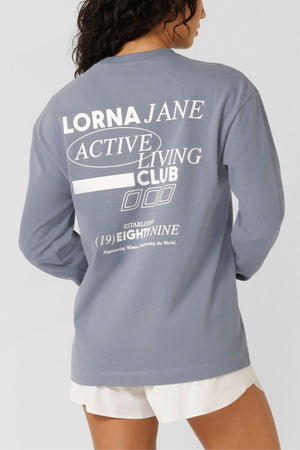 A woman with long, wavy hair faces away from the camera, wearing a light blue Lorna Jane Momentum Plush Long Sleeve T-Shirt with "Lorna Jane Active Living Club" graphics on the back and paired with white shorts.