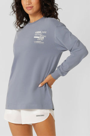A woman wears the Lorna Jane Momentum Plush Long Sleeve T-Shirt in light blue with white chest text, paired with white shorts. She poses with one hand on her hip and smiles slightly against a plain background.
