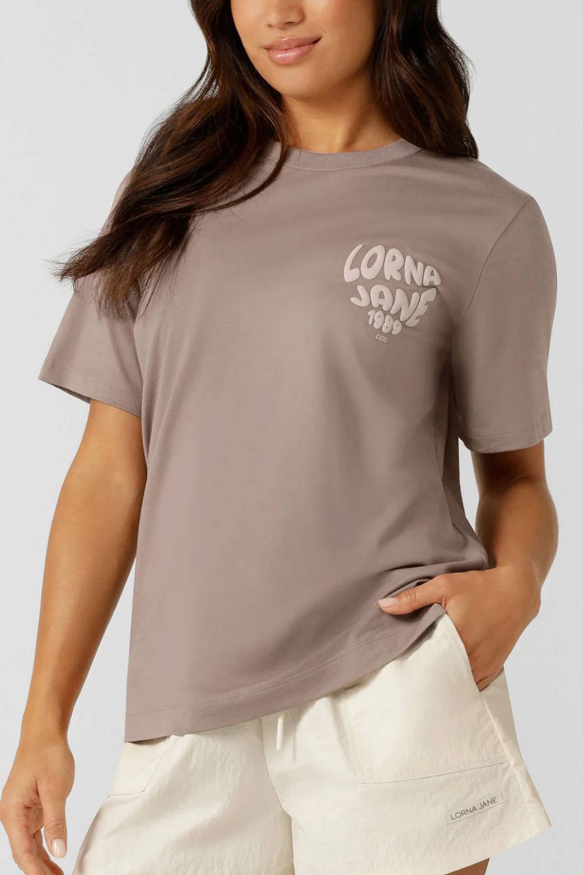 A woman wears the taupe Lorna Jane Never Give Up Relaxed T-Shirt, featuring "Lorna Jane 1990" on the chest, made from a breathable cotton blend, paired with cream shorts, standing with one hand in her pocket against a plain background.