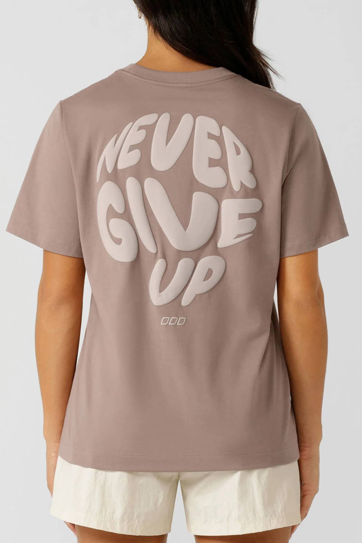 A person wears the limited edition Lorna Jane Never Give Up Relaxed T-Shirt in light brown, featuring bold "NEVER GIVE UP" text and small "OOD" on the back, paired with white shorts against a plain background.