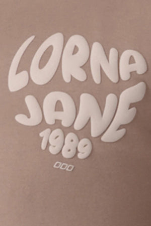 The Lorna Jane Never Give Up Relaxed T-Shirt features "LORNA JANE 1989" in bold, rounded white letters on a light brown background with three small white ovals below the numbers—ideal for your favorite laid-back look.