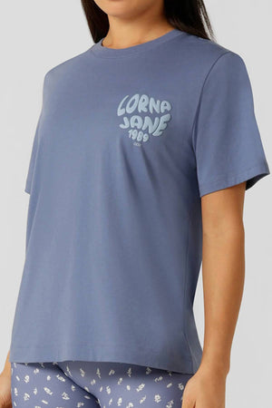 A woman wears a relaxed fit, short-sleeve Lorna Jane Never Give Up Relaxed T-Shirt in light purple with “LORNA JANE 1989” on the chest, paired with printed leggings. Her head is partially out of frame. Perfect for active living.