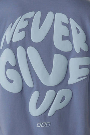A close-up of the Lorna Jane Never Give Up Relaxed T-Shirt in blue, featuring “NEVER GIVE UP” in large, rounded light blue letters on the back; ideal for active living with a relaxed fit.