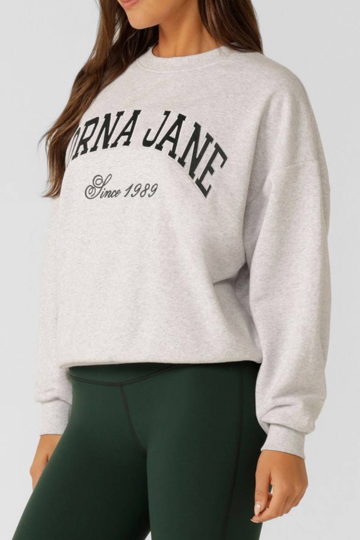 A woman is shown from the waist up wearing the Lorna Jane New Sport Sweatshirt in light grey, featuring "LORNA JANE Since 1989" in black on the front, made from soft cotton terry and styled with dark green leggings.