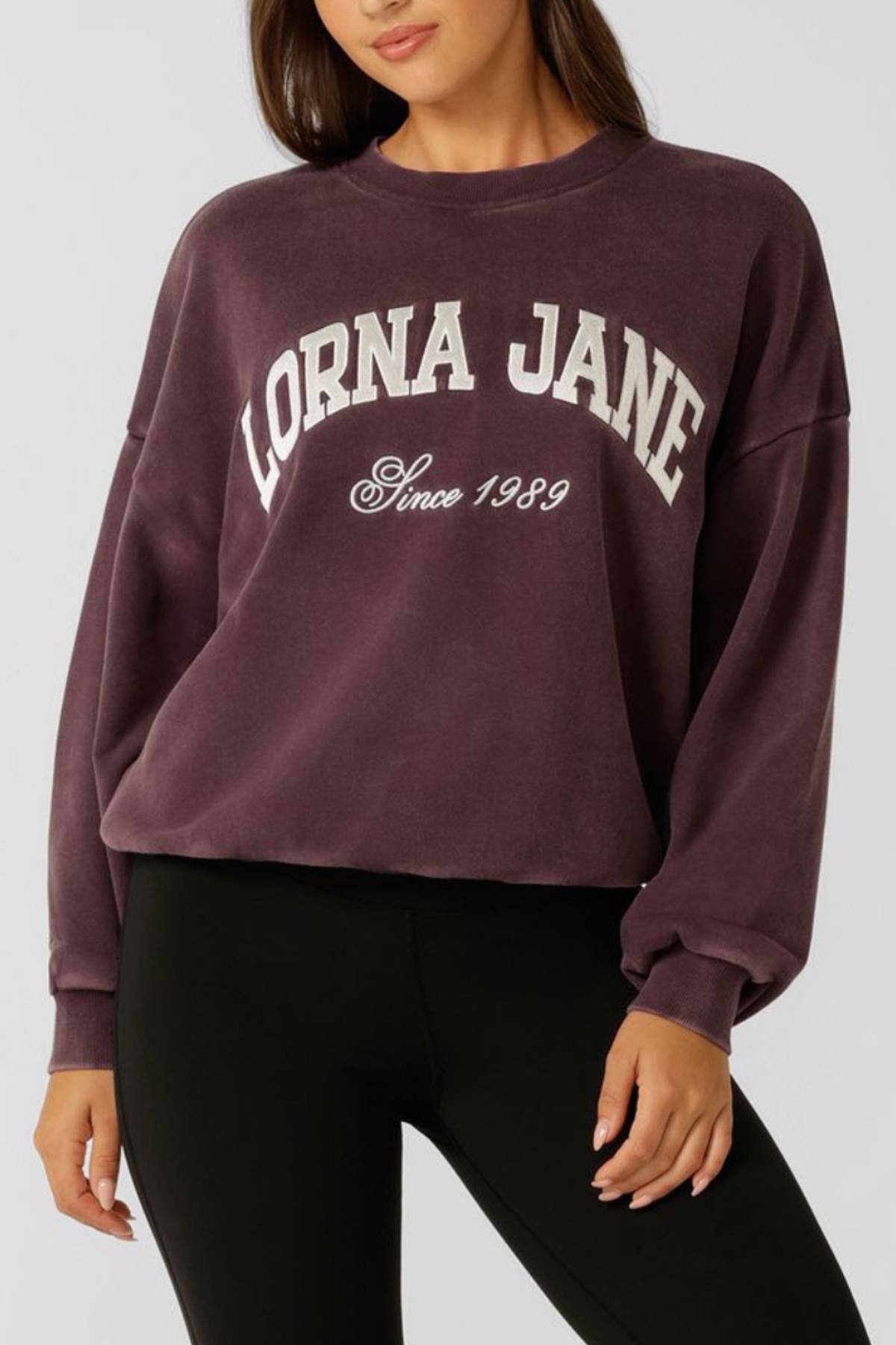 A woman wears a purple Lorna Jane New Sport Washed Sweatshirt with "LORNA JANE Since 1989" in white, paired with black leggings. Her face is partially out of frame.