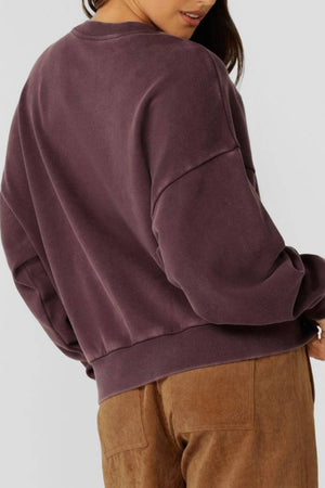A person is seen from behind wearing the Lorna Jane New Sport Washed Sweatshirt in dark mauve with light brown corduroy pants. The plain background highlights the relaxed, casual vibe of their outfit.