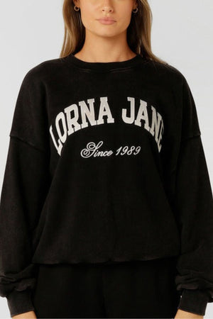 A woman wears the Lorna Jane New Sport Washed Sweatshirt—an oversized, vintage-inspired black cotton terry pullover with "LORNA JANE" and "Since 1989" printed in white. A simple backdrop highlights this classic sporty design.