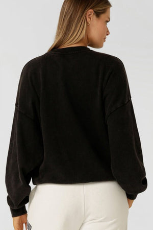 A woman with long, light brown hair faces away from the camera, showing the back of the Lorna Jane New Sport Washed Sweatshirt in an oversized black fit, paired with white pants against a plain light background.