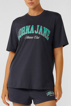 A woman wears the Lorna Jane Original Sport Relaxed T-Shirt in navy blue with "Lorna Jane Athletic Club" in green and white, paired with matching shorts for a sporty, casual, and comfortable everyday look.