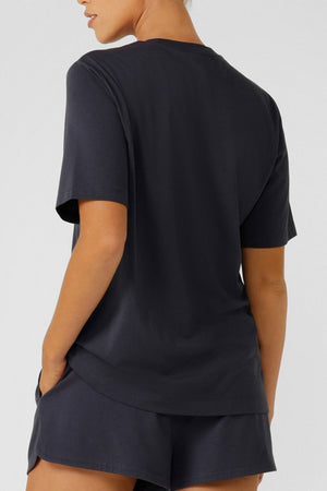 Someone stands with their back to the camera against a plain light background, wearing a Lorna Jane Original Sport Relaxed T-Shirt and matching black shorts.